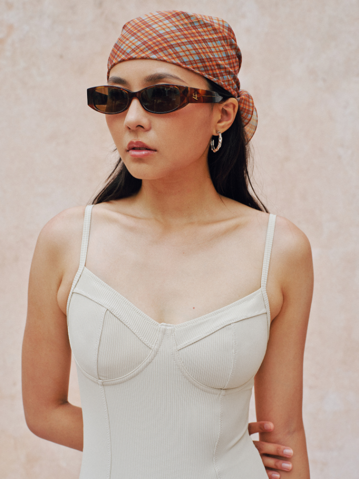 Woman wearing a beige tank top, sunglasses, and a plaid headscarf against a plain background